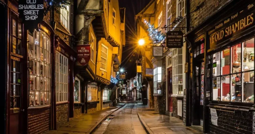 A narrow street at night, featuring shops and buildings, captures the charm of The Shambles illuminated by soft lights.